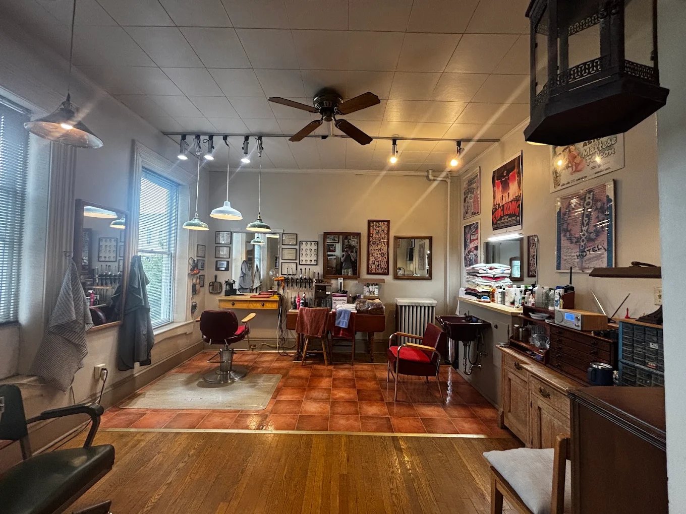 Inside the shop: a red barber chair on a terracotta tile floor, pendant lamps overhead, framed mirrors, and old movie posters on the wall.